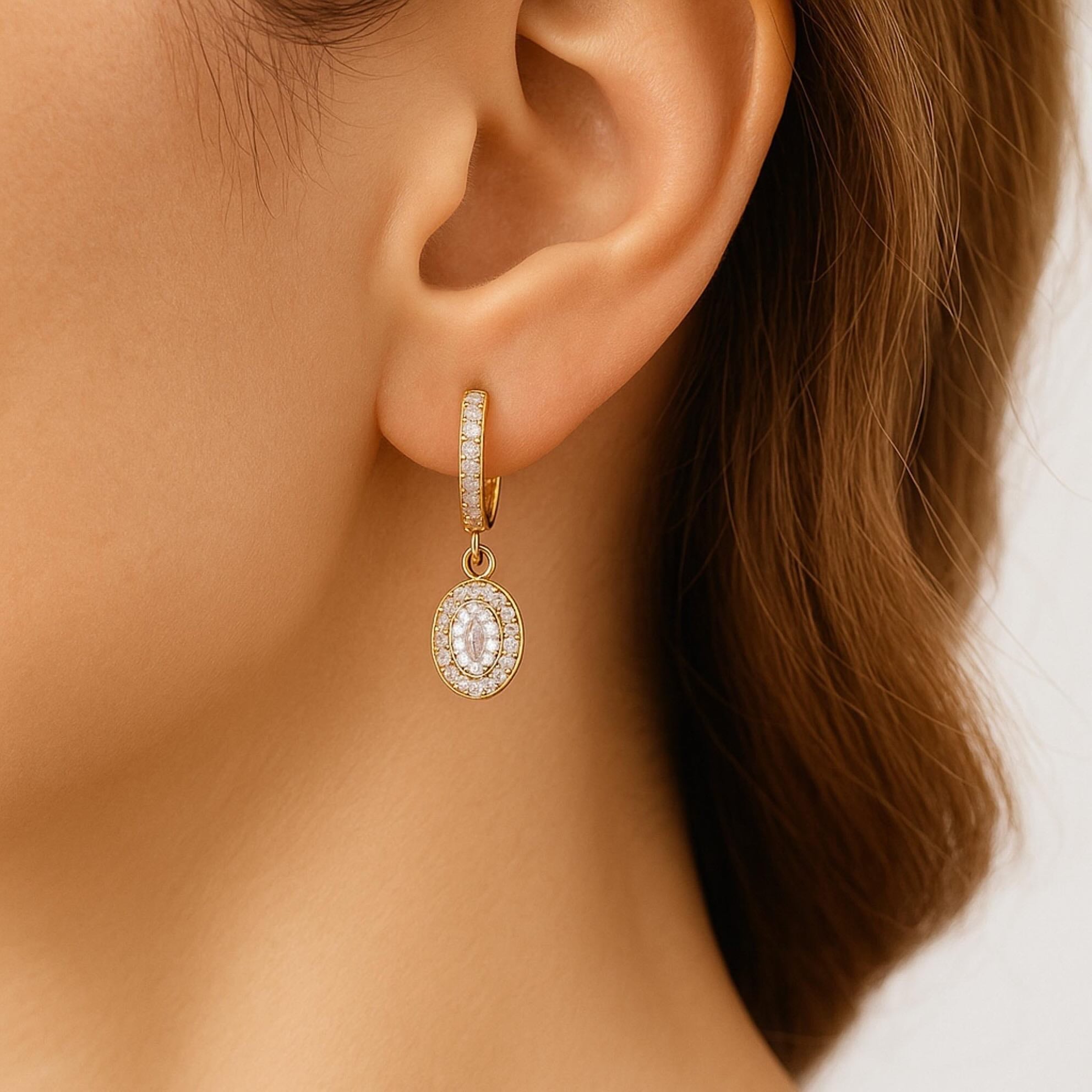 Arete mediano en oro 14K con cierre catalán, diseño colgante de circonia ovalada rodeada de halo brillante, mostrado en la oreja para apreciar su tamaño y elegancia.