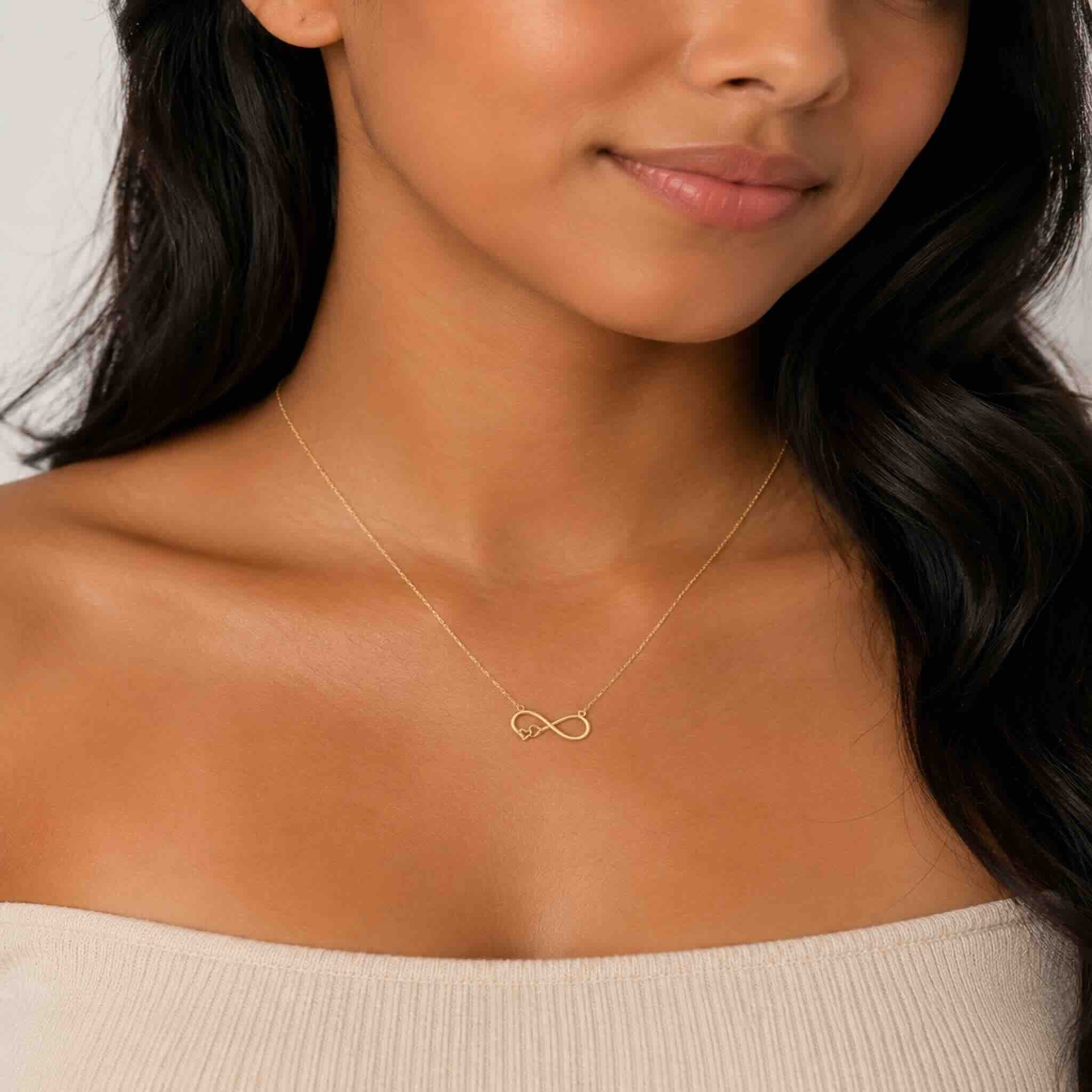 Mujer joven trigueña luciendo cadena fina de oro 10K TuOro con dije de infinito y pequeño corazón, en toma de cuello y escote.