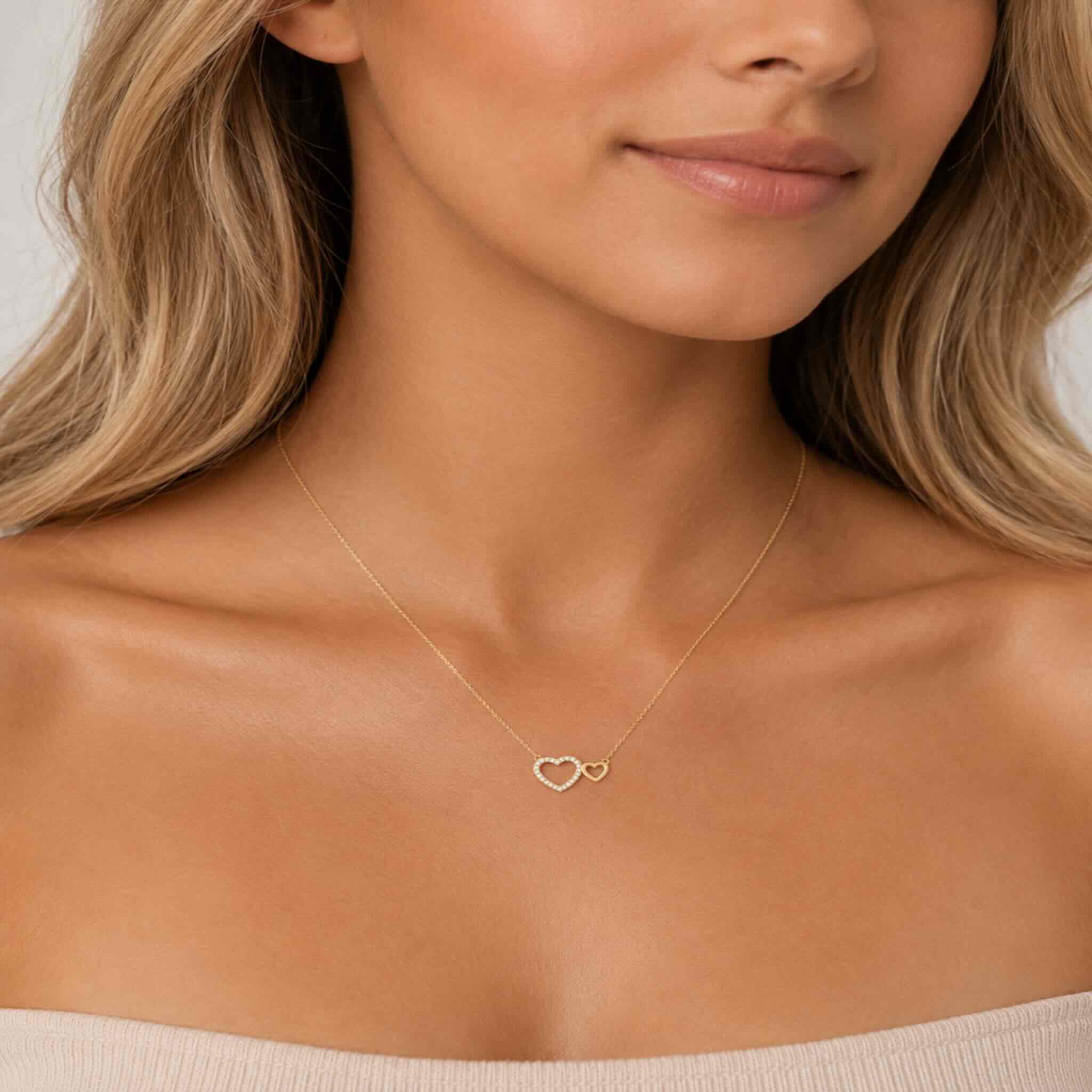 Mujer joven rubia luciendo cadena fina de oro 10K TuOro con dijes de doble corazón, uno con brillo decorativo, en primer plano del cuello.